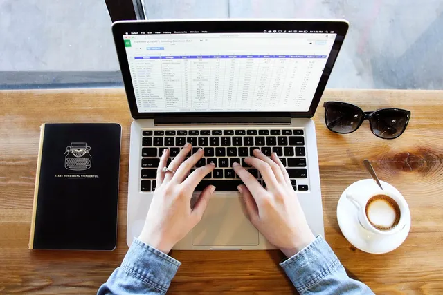 Overhead view of hands typing on a laptop showing a spreadsheet, beside a black notebook, sunglasses and a cappuccino