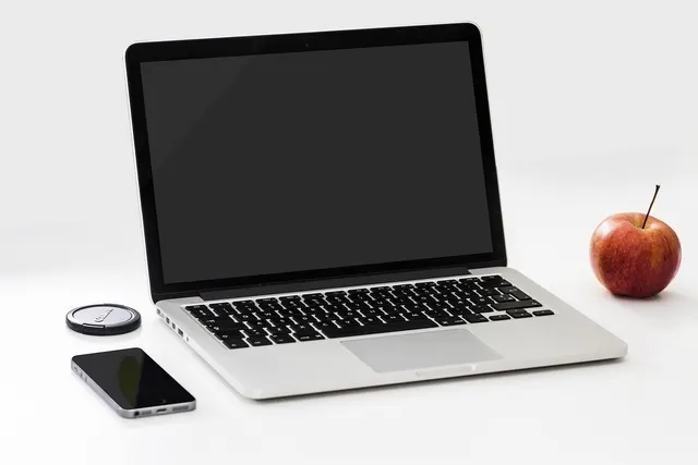 Silver laptop with a blank black screen beside a smartphone, a lens cap and a red apple on a white desk