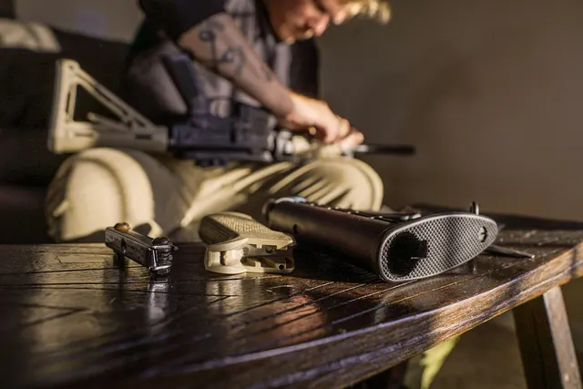 Person assembling an AR-style rifle on a couch, with a pistol grip, buttstock and small part on a wooden table in front