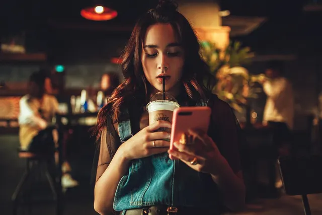 Young woman sipping an iced drink while looking at her pink smartphone in a dimly lit cafe
