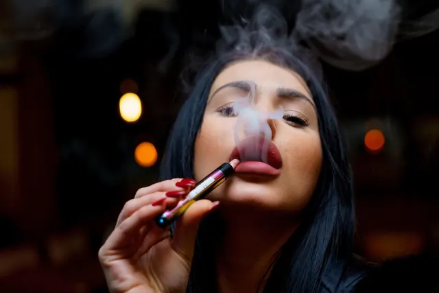 Dark-haired woman with red nails exhaling vapor from a colorful disposable vape pen against blurred warm lights