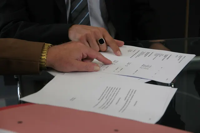 Two businesspeople in suits pointing at lines on printed documents spread across a glass table beside a pink folder