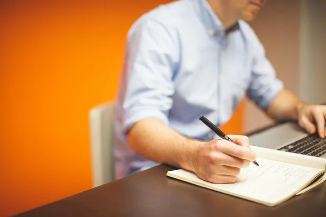 Man in a light blue shirt writing notes in a notebook while typing on a laptop at a desk against an orange wall