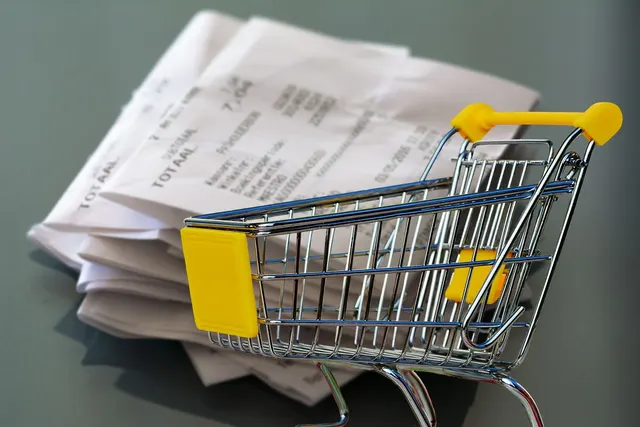Miniature metal shopping cart with yellow handle in front of a pile of crumpled printed receipts on a gray surface