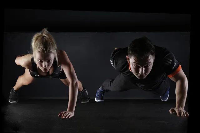 Woman and man in black workout clothes doing one-arm push-ups side by side on a dark gym floor