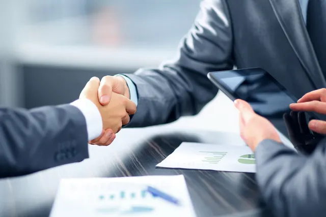 Two businesspeople in suits shake hands over a desk with financial chart printouts as a third person holds a tablet