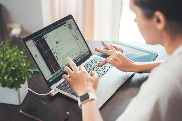 Woman typing on a laptop at a wooden desk, wearing a smartwatch, with a smartphone and potted plant beside her
