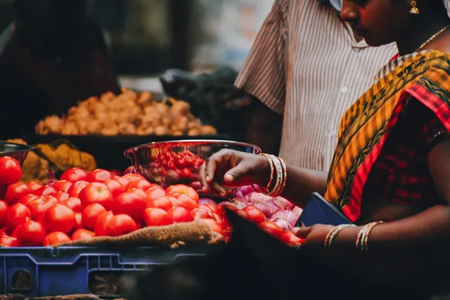 Woman in a sari and gold bangles holding a blue wallet at a market stall with tomatoes, onions and potatoes