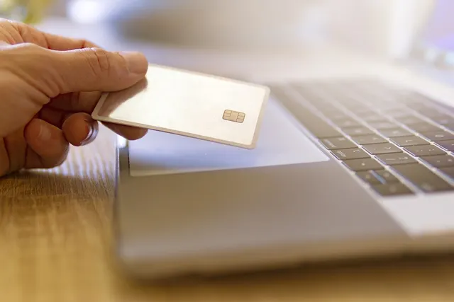 Hand holding a blank gold chip credit card above a silver laptop keyboard on a wooden desk, suggesting an online payment