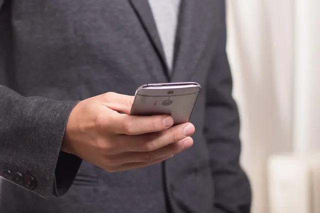 Close-up of a man in a grey blazer holding a silver smartphone, viewing the screen in a bright room