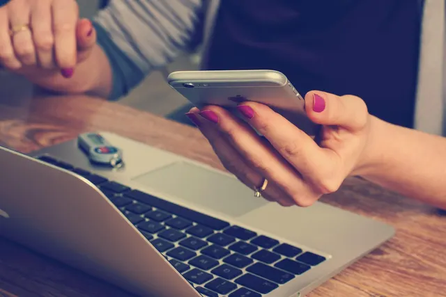 Woman with pink nails holding a smartphone above an open laptop with a USB key resting on it at a wooden desk