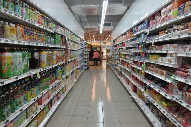Empty grocery store aisle with shelves of bottled drinks on the left and packaged snacks on the right, a shopper at the far end