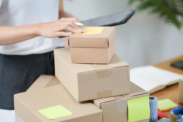 Woman holding a tablet and pen places a yellow sticky note on a stack of taped cardboard shipping boxes on a desk