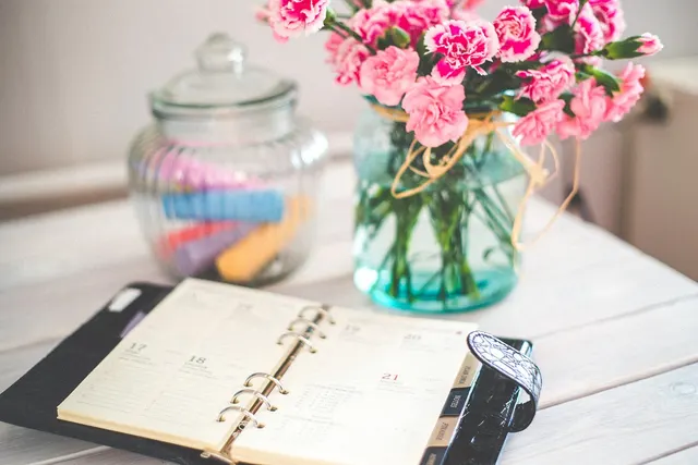 Open ring-bound planner showing dated pages on a white wooden desk beside pink carnations and a jar of colored chalk