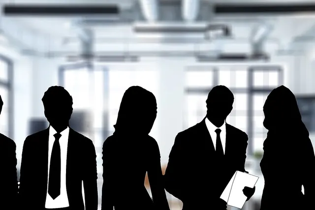 Black silhouettes of six businesspeople in suits, one holding papers, against a blurred bright modern office