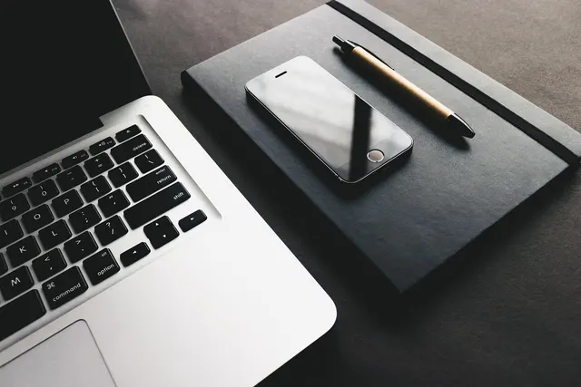 Silver laptop keyboard beside a black smartphone and pen resting on a dark notebook on a gray desk