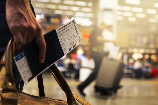 Traveler holding a passport and boarding passes beside a leather bag in a busy airport terminal
