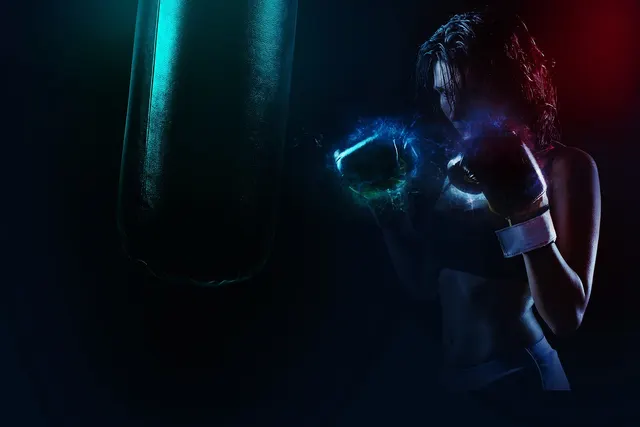 Woman in boxing gloves throwing a punch at a hanging heavy bag in a dark gym lit with teal and red light