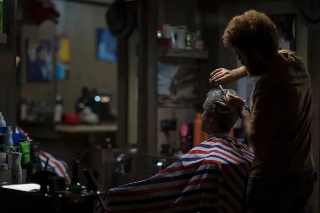 Barber trimming an older man's gray hair with scissors in a dimly lit barbershop, client draped in a striped cape