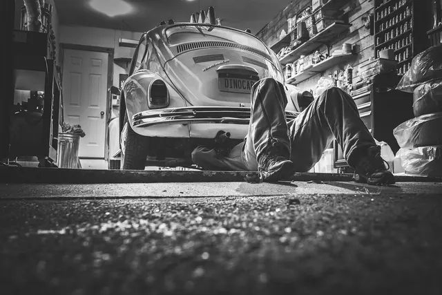 Mechanic lying on his back under a classic beetle-style car in a cluttered home garage, black and white