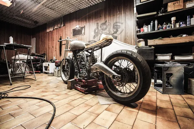 Custom motorcycle on a lift stand in a wood-paneled repair garage with tool shelves and a workbench
