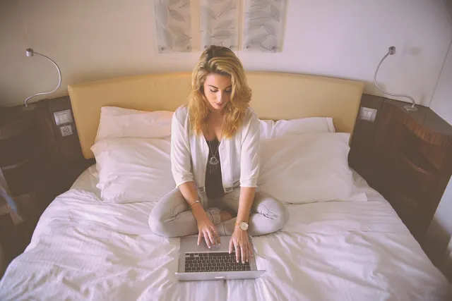 Woman sitting cross-legged on a white hotel bed typing on a laptop, with bedside lamps and leaf prints behind her