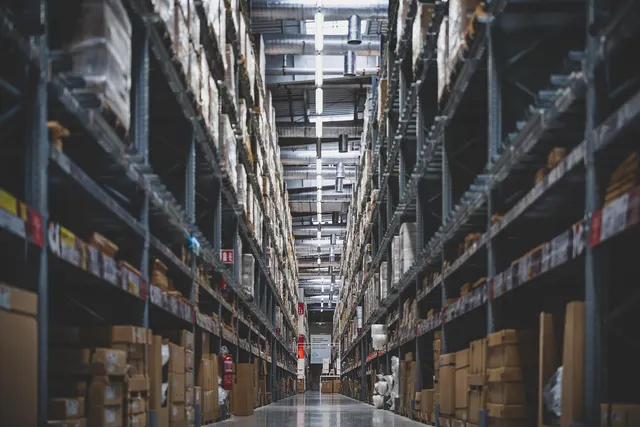 Long warehouse aisle with tall metal shelving stacked with cardboard boxes and pallets under overhead lights