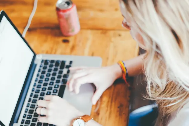 Overhead view of a blonde woman typing on a laptop at a wooden table, wearing a watch, with a soda can nearby