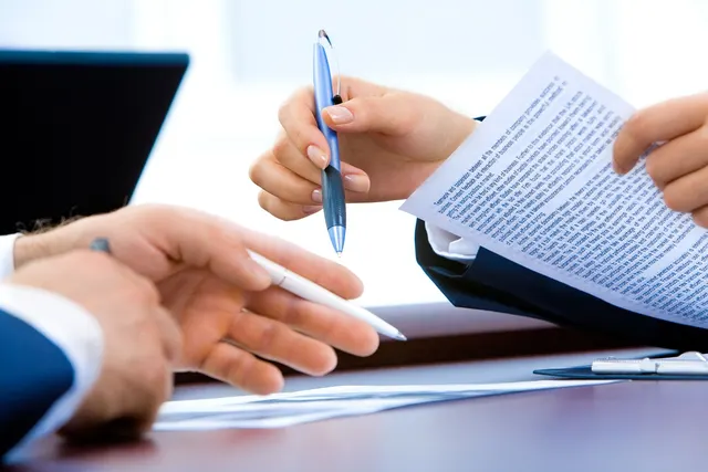 Two professionals in suits holding pens while reviewing a printed document across a desk with a laptop