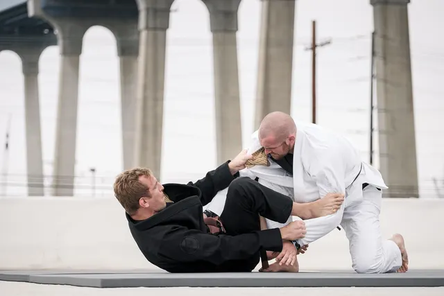 Two men in black and white jiu-jitsu gis grappling on a mat outdoors beneath a concrete bridge overpass