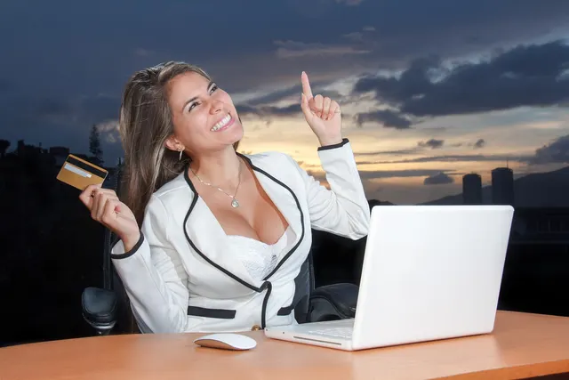 Smiling woman in white blazer holds a gold credit card and points upward while seated at a desk with a laptop at dusk