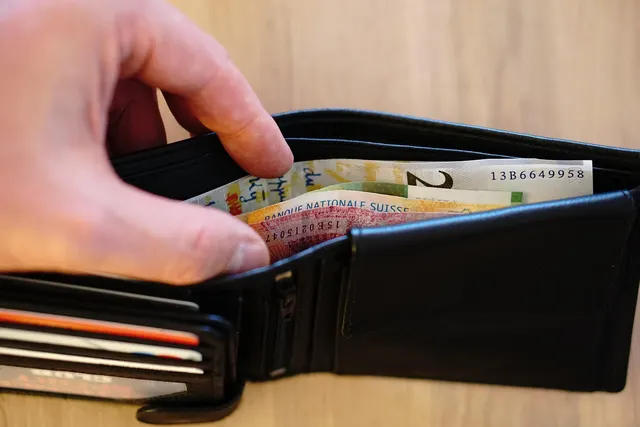 Hand opening a black leather wallet holding Swiss franc banknotes and payment cards on a wooden table