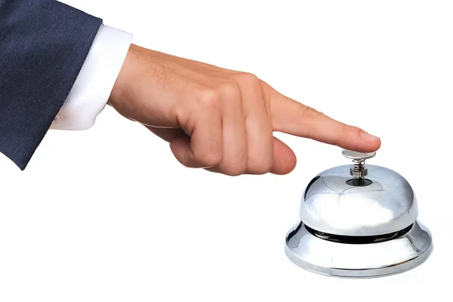 Hand in a navy suit sleeve pressing a silver front desk service bell on a white background
