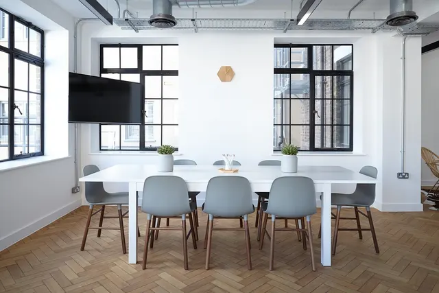 Bright, empty meeting room with a white table, grey chairs, potted succulents, a wall-mounted TV and black-framed windows