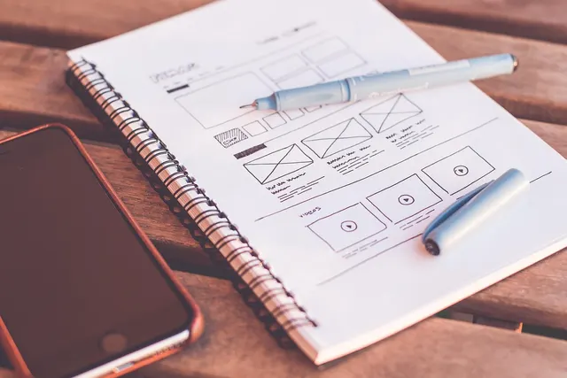 Spiral notebook with hand-drawn website wireframe sketches, a light blue pen and a smartphone on a wooden table