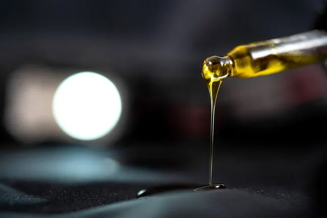Close-up of a glass dropper releasing a thin stream of golden CBD oil onto a dark surface with a blurred light behind