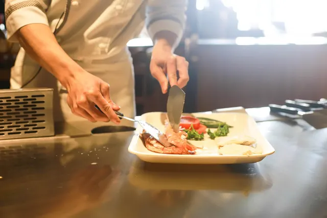 Chef in white jacket using a spatula and knife to plate grilled shrimp and vegetables on a teppanyaki grill counter