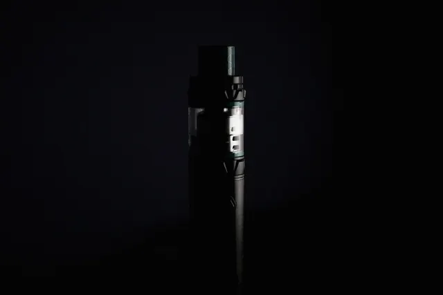 Black vape pen with clear tank standing upright against a dark background, lit by a narrow side light