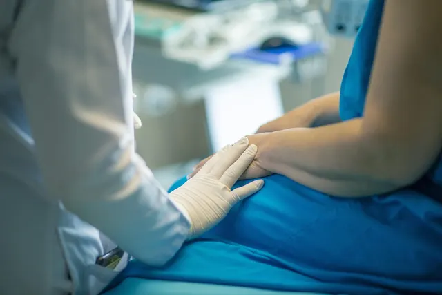 Clinician in white coat and latex gloves rests a hand on a patient's hands as she sits in blue scrubs in an exam room
