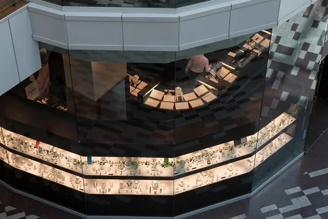 Overhead view of a curved glass jewelry kiosk in a mall, its lit display cases lined with necklaces and rings