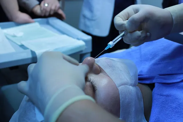 Gloved provider injects a syringe near the nose of a patient whose head is wrapped in gauze on a blue-draped treatment table