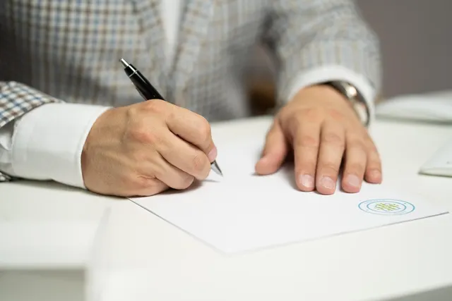 Close-up of a man in a checked blazer signing a document with a black pen at a white desk
