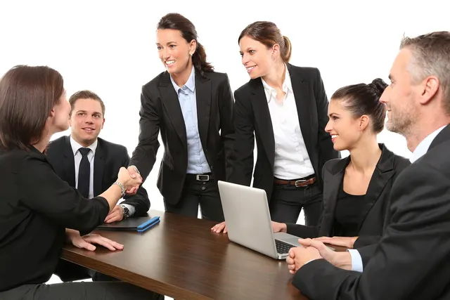 Two businesswomen shake hands across a meeting table as colleagues in dark suits smile, a laptop open on the table