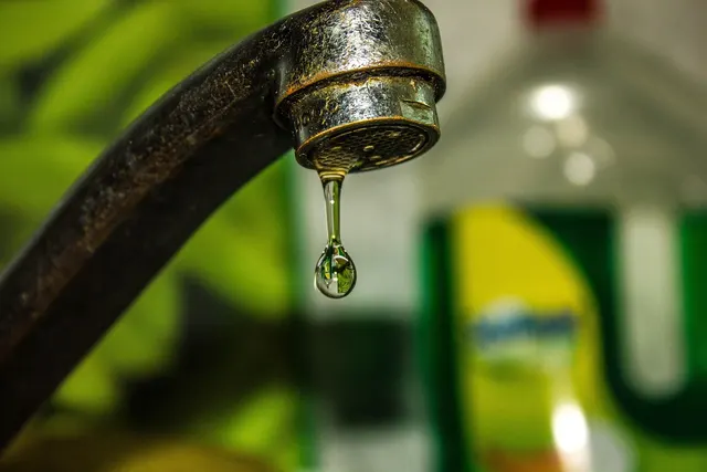 Close-up of a worn, corroded kitchen faucet with a single water drop falling from the spout against a blurred background