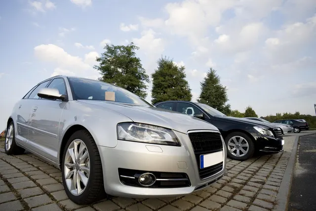 Silver hatchback and black sedan parked on a cobblestone car lot under a partly cloudy sky with trees behind