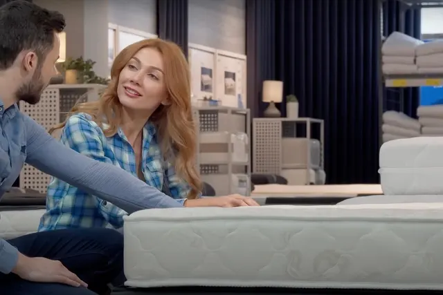 Couple sitting beside a white mattress in a mattress store, testing it before purchase, with stacked mattresses behind