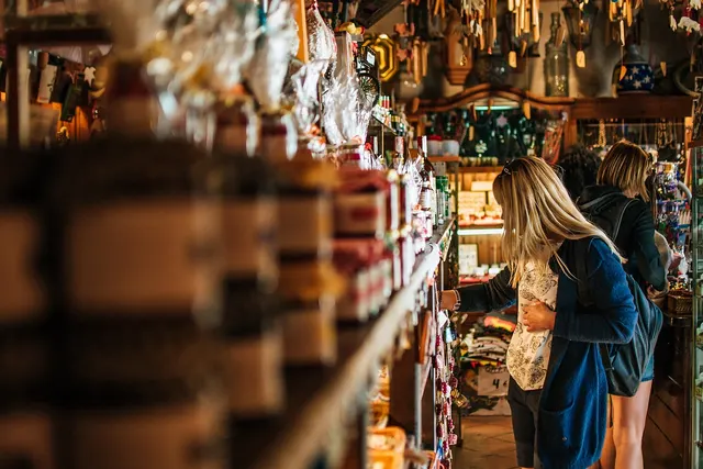 Two women with backpacks browse crowded shelves of jars, bottles and gifts in a small independent retail shop
