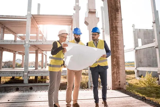 Three workers in hard hats reviewing blueprints at a concrete building site