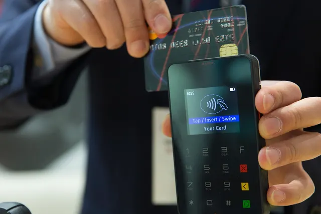 Hand holding a chip credit card above a handheld card reader whose screen reads Tap / Insert / Swipe Your Card