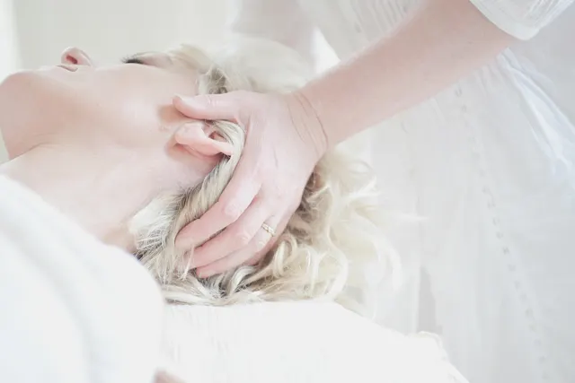 Massage therapist's hand cradling the head of a blonde woman lying relaxed on a massage table in a bright white room
